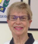 An older woman with short hair and glasses smiles broadly, wearing a karate uniform, against a backdrop of martial arts certificates.