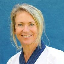 A smiling woman wearing a martial arts uniform, with light blonde hair and a blue background.