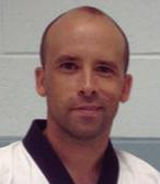 A man with a bald head, wearing a martial arts uniform, smiles against a light blue background.