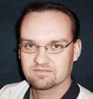 A man with glasses and short hair, showing a serious expression against a dark background.