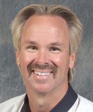 A smiling man with light blonde hair and a mustache, wearing a black and white outfit, set against a gray background.