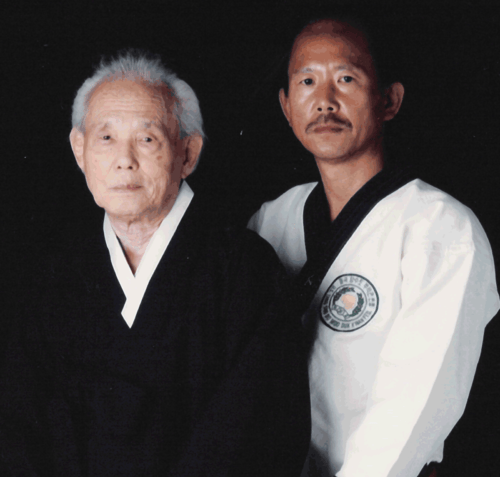 Two men in traditional attire, one elderly and the other middle-aged, positioned closely against a dark backdrop.