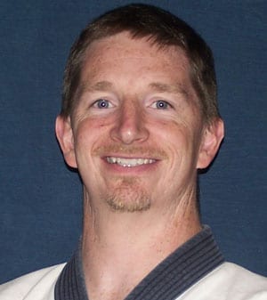 A smiling man in a martial arts uniform, with short hair and a light goatee, against a blue background.