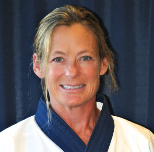 A smiling woman in a martial arts uniform with a dark blue collar, set against a dark background.