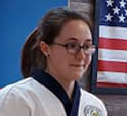 A young female martial artist in a traditional uniform poses confidently, with an American flag visible in the background.