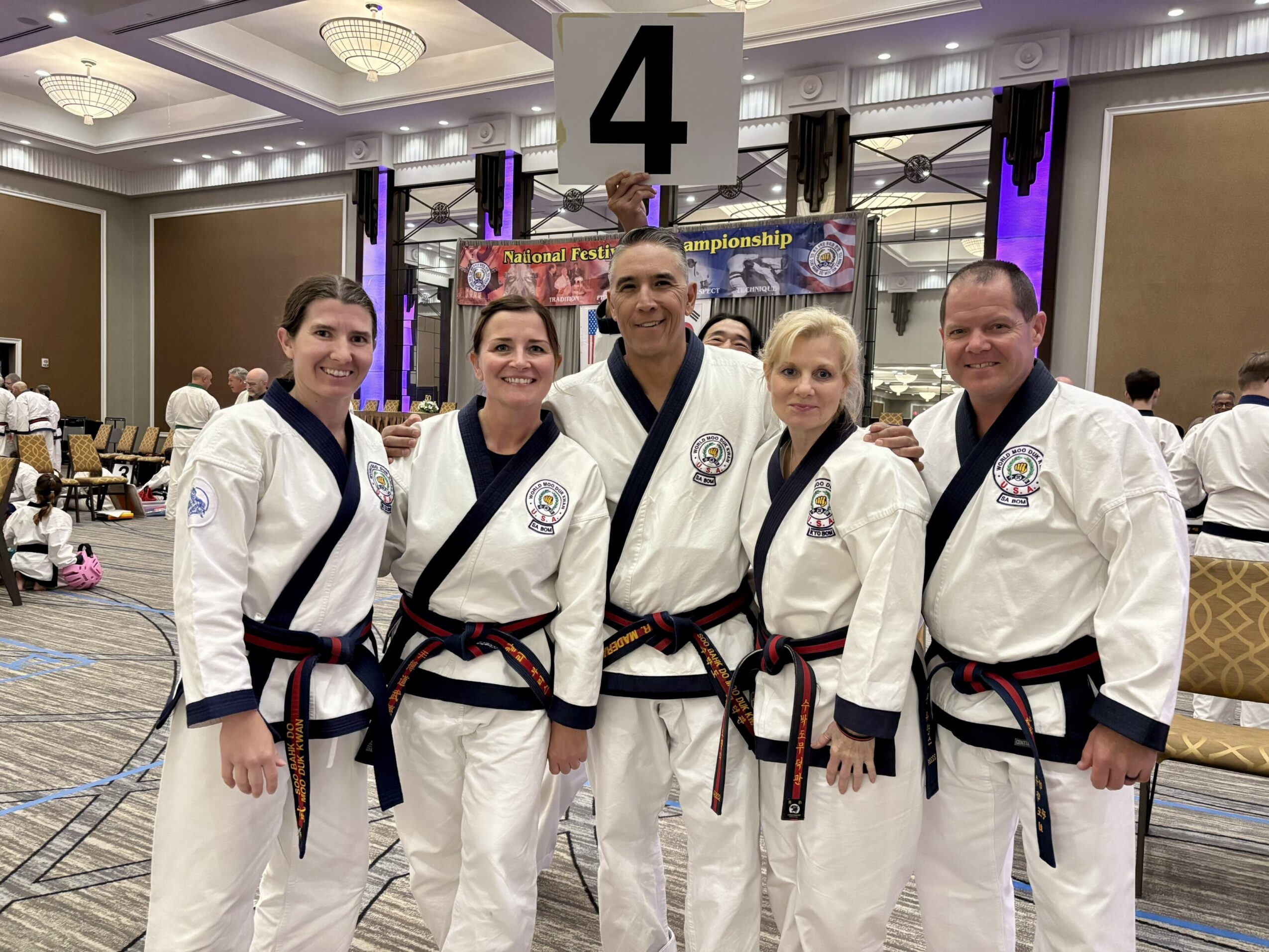 Five martial artists in traditional uniforms pose together, smiling, with a competition banner in the background and a large number four sign above them.